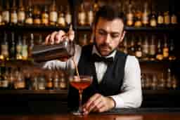 Bartender in a black vest and bow tie preparing a cocktail in a bar with liquor bottles in the background, representing APLH Personal Licence Training Course
